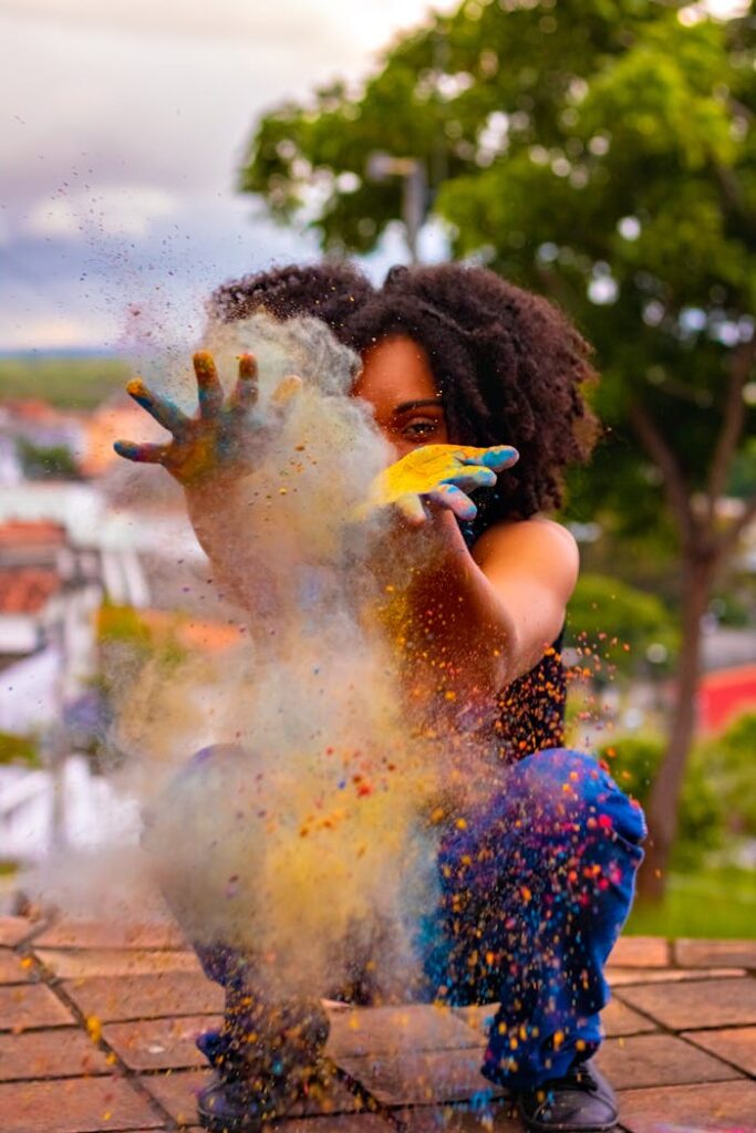 Anonymous African American female squatting on tile pavement with hands apart while playing with multicolored dry paints during Hindu Festival of Colors under cloudy sky